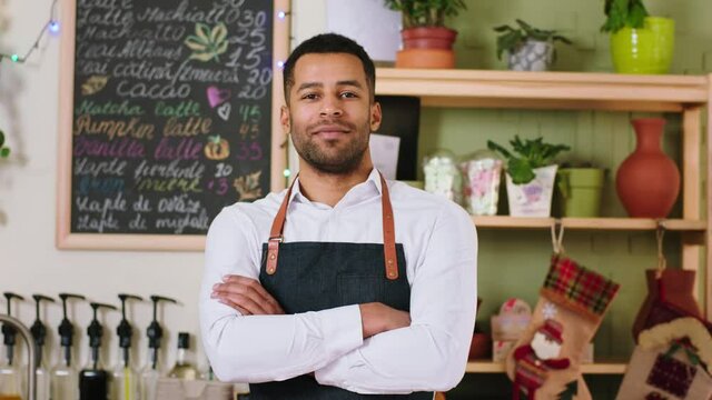 Concept of small business and entrepreneur black guy very charismatic with a large smile at tue coffee shop desk standing and posing in front of the camera crossing hands