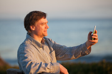 Caucasian man using cellphone relaxing by ocean at sunset