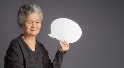 A speech bubble concept. A senior woman holding and looking at a blank bubble balloon while standing on a gray background