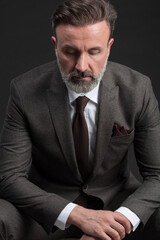 Portrait of adult businessman wearing trendy suit and sitting in modern studio on stylish chair against the black background