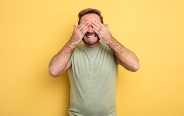 middle age handsome man smiling and feeling happy, covering eyes with both hands and waiting for...