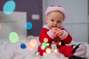 Baby boy wearing in santa claus sits on the bed playing with lights.