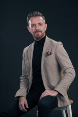 Portrait of adult businessman wearing trendy suit and sitting in modern studio on stylish chair against the black background