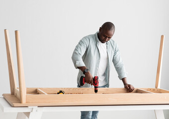 Professional african american craftsman drilling wooden planks of table, assembling new furniture and screwing details
