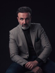 Portrait of adult businessman wearing trendy suit and sitting in modern studio on stylish chair against the black background