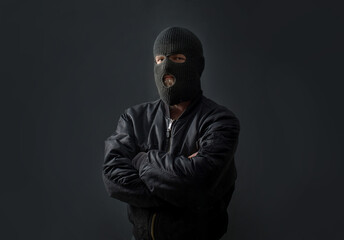 Naklejka na ściany i meble a man stands against a dark background in a black bomber jacket and a balaclava mask on his face