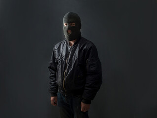 a man stands against a dark background in a black bomber jacket and a balaclava mask on his face