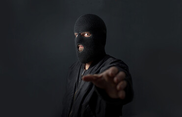 a man stands against a dark background in a black bomber jacket and a balaclava mask on his face...