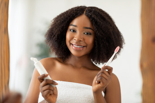 Dental care. Attractive young black woman with radiant smile holding toothpaste and brush in front of mirror at home