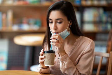 Arab lady drinking coffee with straw in cafe after quarantine, lowered medical mask on her chin and enjoying hot drink