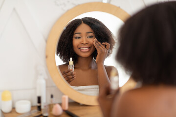 Beauty routine procedures. Sensual young black woman applying hydrating serum in front of mirror at home