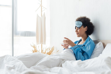 joyful african american woman in pajamas and sleeping mask holding cup of coffee in bed