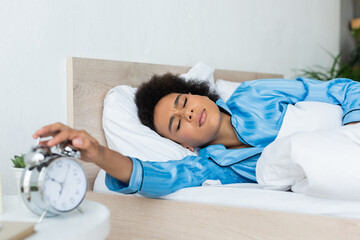 sleepy african american woman in pajamas frowning while reaching alarm clock