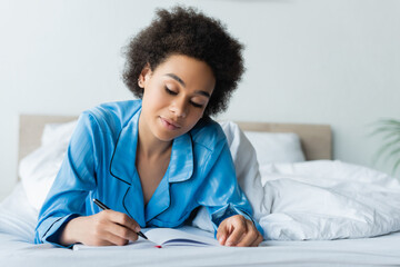 happy african american woman in pajamas lying on bed and writing on notebook
