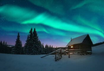 Fantastische Winterlandschaft mit Holzhaus mit Licht im Fenster in schneebedeckten Bergen und Nordlicht im Nachthimmel. Weihnachtsferien und Winterferienkonzept