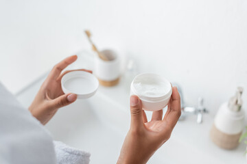 cropped view of african american woman holding container with face cream in bathroom