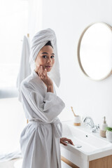 young african american woman in bathrobe and towel applying face cream in bathroom