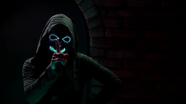 threatening person with glowing skull mask and black hood is making forbidding gesture