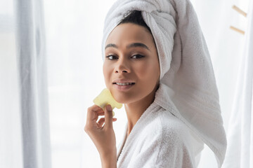 pretty african american woman in white towel holding jade face scraper near face