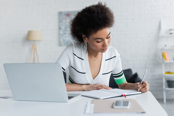 african american woman writing on notebook near gadgets