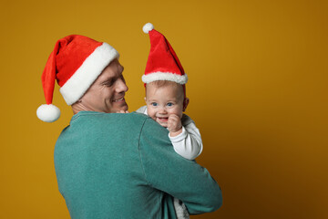 Happy father with cute baby wearing Santa hats on yellow background. Christmas season