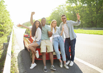 Portrait of happy diverse friends posing near their car on highway, making different gestures, smiling at camera