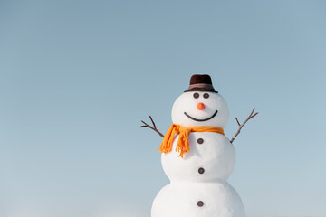 Funny snowman in stylish brown hat and yellow scalf on snowy field. Blue sky on background