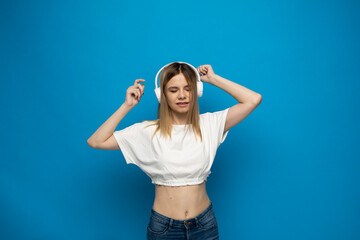 Fototapeta na wymiar Beautiful attractive young blond woman wearing white t-shirt and glasses in white headphones listening music and dancing on blue background in studio. Relaxing and enjoying. Lifestyle.