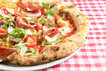 Cropped photo of a baked pizza with prosciutto, ham Iberian, buffalo mozzarella, cheese. Traditional Italian dish on red and white checkered tablecloth. Close-up, copy space, top view.