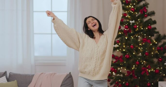 Funny girl dancing, having fun while celebrating winter holidays. A happy festive woman celebrating New Year or Xmas near decorated Christmas tree indoors