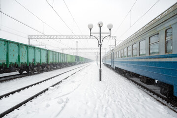 Train station peron covered by snow in winter time. Passenger train and freight wagons on snowy...