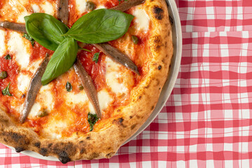 Cropped photo of a baked pizza with buffalo mozzarella, parmigiano-reggiano cheese, basil and anchovies. Traditional Italian dish on red and white checkered tablecloth. 
