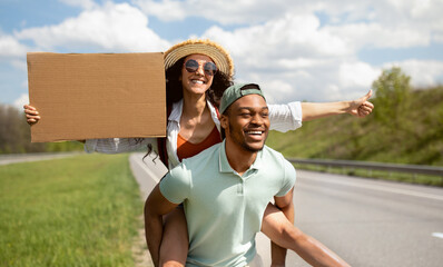 Young black guy giving piggyback ride to his girlfriend, hitchhiking on road together, holding...