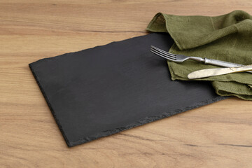 Black slate board and fork,knife and green napkin on the wooden table.Free space