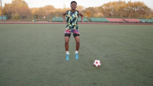 Young black girl warming up in training on the football field next to the ball and jumping vigorously raising her legs high