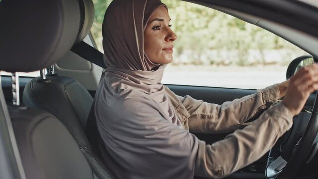 Tilt up shot of Muslim woman in hijab sitting in car and putting seat belt on before driving