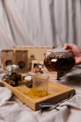 A hand pouring tea from a glass teapot onto a wooden serving tray. A jar of tea and a candle on a textile background.