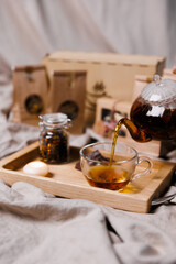 A hand pouring tea from a glass teapot onto a wooden serving tray. A jar of tea and a candle on a textile background.
