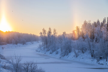 Beautiful winter landscape. Sunrise on a frozen river. The rising sun is like a candle, and a rainbow is visible on the other side of the sky. Trees are covered with hoarfrost 