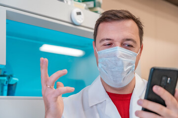 Fototapeta premium Excited scientist wearing white coat showing emotions of joy about victory in online lottery in room with laboratory box in UV ligh. Amazed male in medical mask win money celebration success