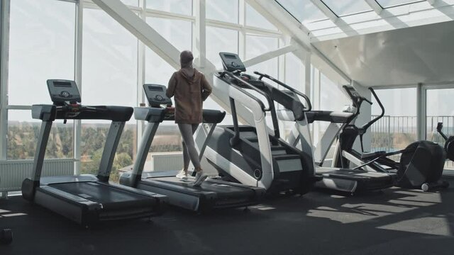 Back View Wide Shot Of Woman In Hijab And Sportswear Jogging On Treadmill In Modern Gym