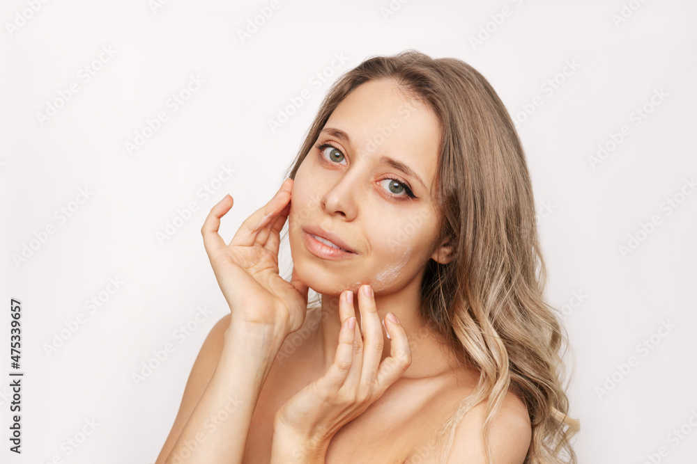 Wall mural A young beautiful caucasian woman with wavy hair touching the face with her hands applying a moisturizing cream on the skin isolated on a white background. Cosmetology and beauty concept