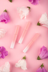 Lip gloss in a pink-to-white gradient cases with white and purple hibiscus flowers on a pink background. Flat lay trending scene with buds. Frame made of flowers. Decorative cosmetics liquid lipstick