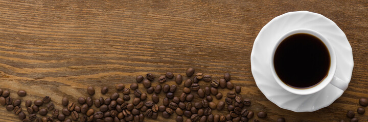 White cup of black coffee with brown beans on dark wooden table background. Closeup. Wide banner. Empty place for text. Top down view.