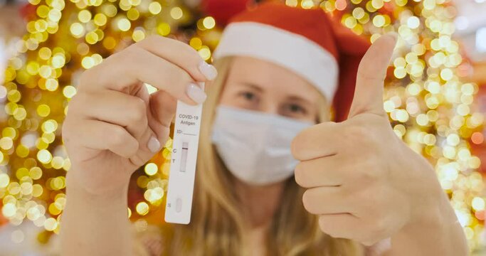 Girl shows negative COVID-19 antigen test and thumbs up sign. Woman with face mask and Christmas hat of Santa Claus stands by tree with lights and garlands decorated for New Year, no logo