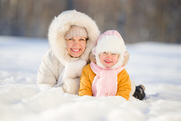 Fototapeta na wymiar Happy young mother and little daughter smiling and lying down on white snow at nature park. Lovely emotional moment. Spending time together in beautiful cold sunny winter day. Front view. Closeup.