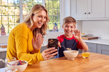 Mother And Son At Home Eating Breakfast Cereal At Kitchen Counter And Making Video Call On Mobile Phone