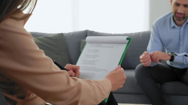 Close up back view woman psychologist taking notes on clipboard and listens patient at psychotherapy session. Unfocused adult man with mental health problems sitting on couch talking to therapist