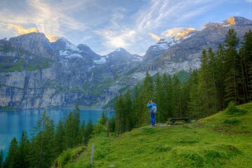 Fototapeta na wymiar Tourist enjoying the scenery of peaceful Oeschinen Lake with a wooden farm house by the lakeside & majestic mountains in background on a beautiful summer morning in Bernese Oberland, Switzerland