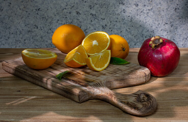 Still life: a group of fresh oranges, whole and cut, lies on an old figured wooden board and a ripe pomegranate next to it.  Drawing with light and shadow. Selective focus